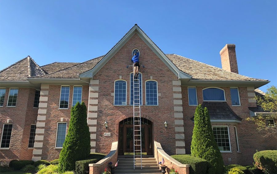 Professional window washer on a ladder cleaning a brick home — the benefits of clean windows start outside